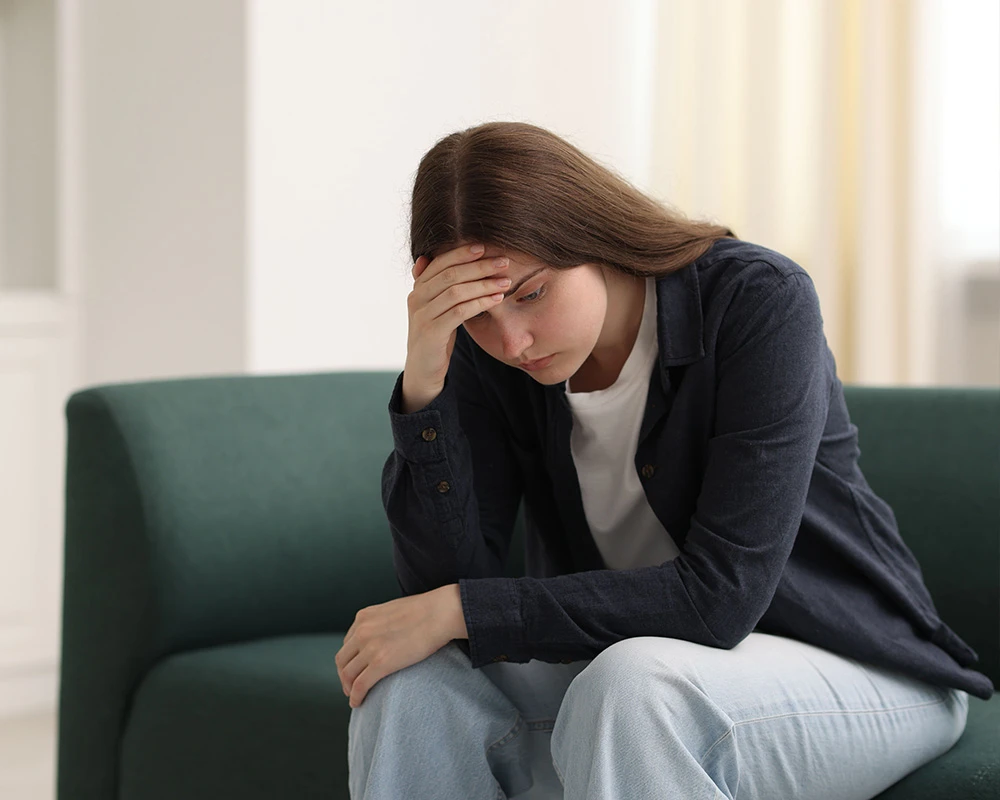 Jonge vrouw zit bezorgd op de bank met haar hand tegen haar hoofd. De afbeelding past bij emotionele en mentale symptomen van gokverslaving.