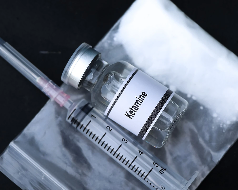 Flesje met ketamine ligt naast een injectiespuit en een zakje met wit poeder. De afbeelding verwijst naar ketaminegebruik en verslavingsrisico’s.