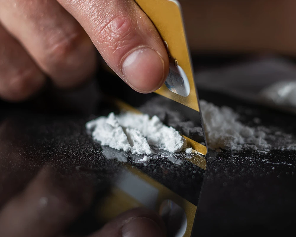 Persoon verdeelt wit poeder met een scherp voorwerp op een donkere ondergrond. De afbeelding past bij informatie over 3MMC verslaving en drugsgebruik.