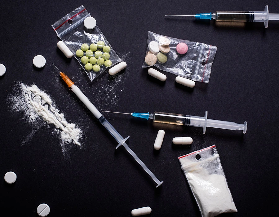 Zakjes met wit poeder, pillen en injectiespuiten liggen verspreid op een donkere ondergrond. De afbeelding verwijst naar verschillende middelen die bekendstaan als de meest verslavende drugs.