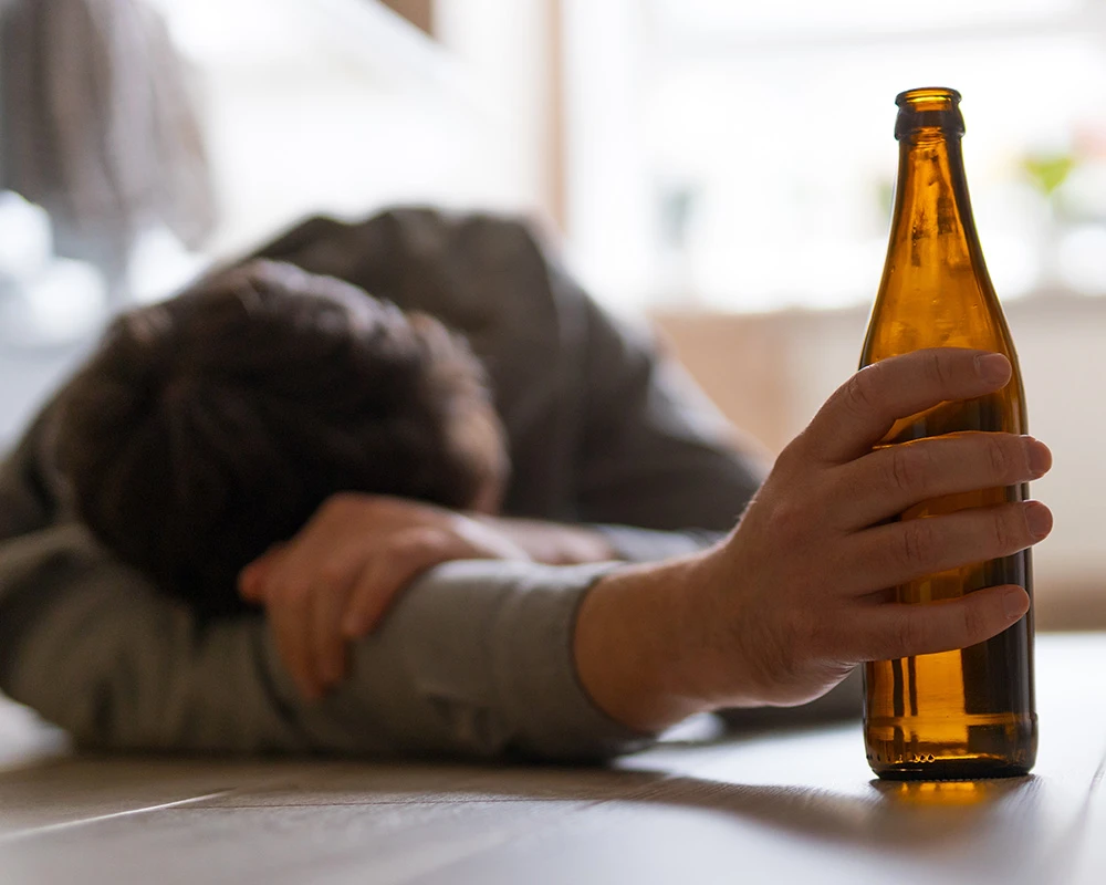 Persoon ligt op de grond en reikt naar een fles alcohol. De afbeelding past bij signalen en gedrag die kunnen wijzen op een terugval in verslaving.