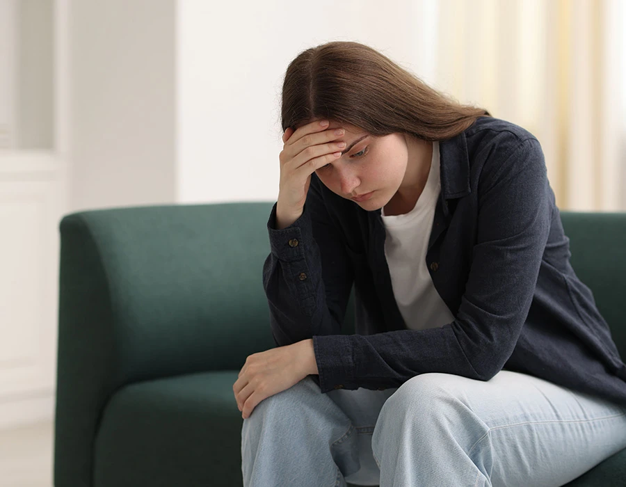 Een jonge vrouw zit bezorgd op de bank met haar hand tegen haar hoofd en denkt na over mogelijke verslavingsproblemen en mentale klachten.