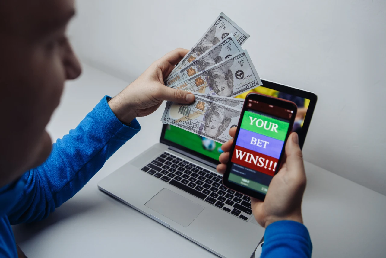 Man voedt online gokverslaving door op smartphone en laptop te gokken, waarbij hij geld vasthoudt.