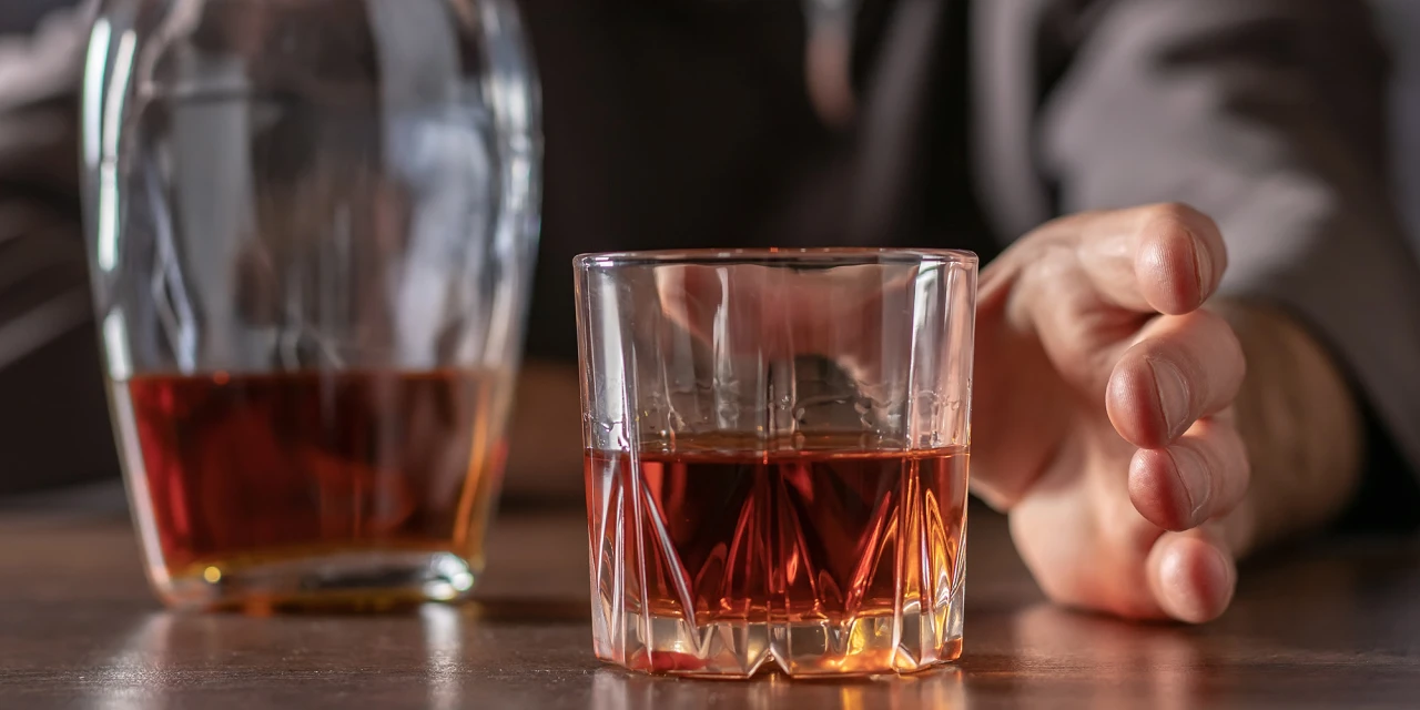 Een persoon reikt naar een glas alcohol terwijl een fles sterke drank op tafel staat tijdens een moment van twijfel of controleverlies.