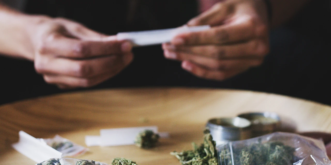 Iemand rolt een joint terwijl cannabis, vloeitjes en accessoires op tafel liggen.
