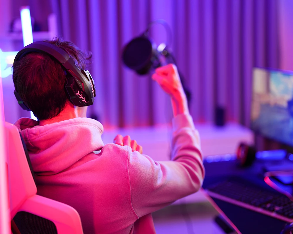 Iemand met een headset zit achter een gaming setup en juicht tijdens het spelen van FIFA. De afbeelding verwijst naar intensief gamen en gedrag dat kan passen bij verslaafd zijn aan FIFA.