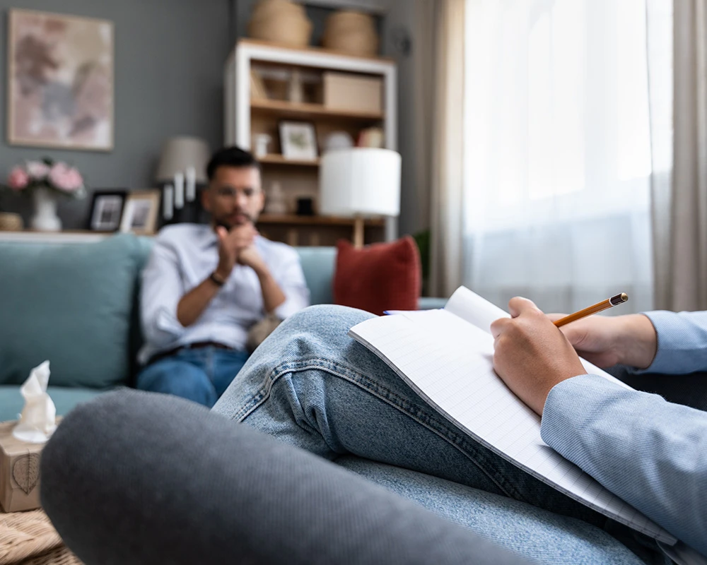 Een behandelaar maakt aantekeningen tijdens een gesprek met iemand op de bank. De afbeelding laat begeleiding en ondersteuning zien die kan passen bij hulp bij gokverslaving.