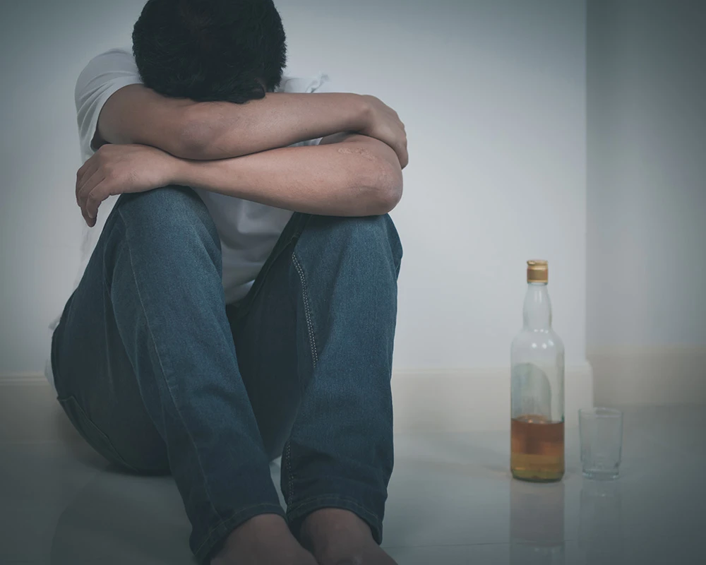 Persoon zit verdrietig op de grond naast een fles alcohol en een glas. De afbeelding past bij informatie over verslaving, mentale klachten en de vraag of verslaving erfelijk is.