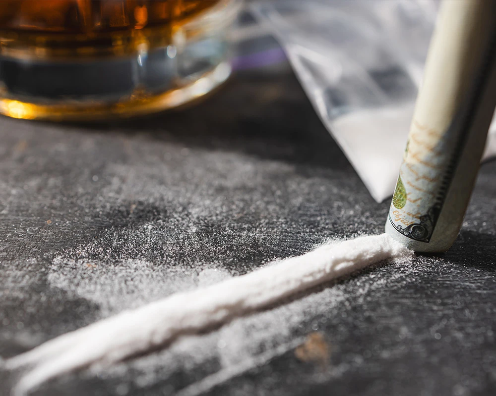 Een lijn cocaïne ligt op tafel naast alcohol en opgerold geld. De afbeelding toont middelengebruik en de risico’s van alcohol en coke combineren.