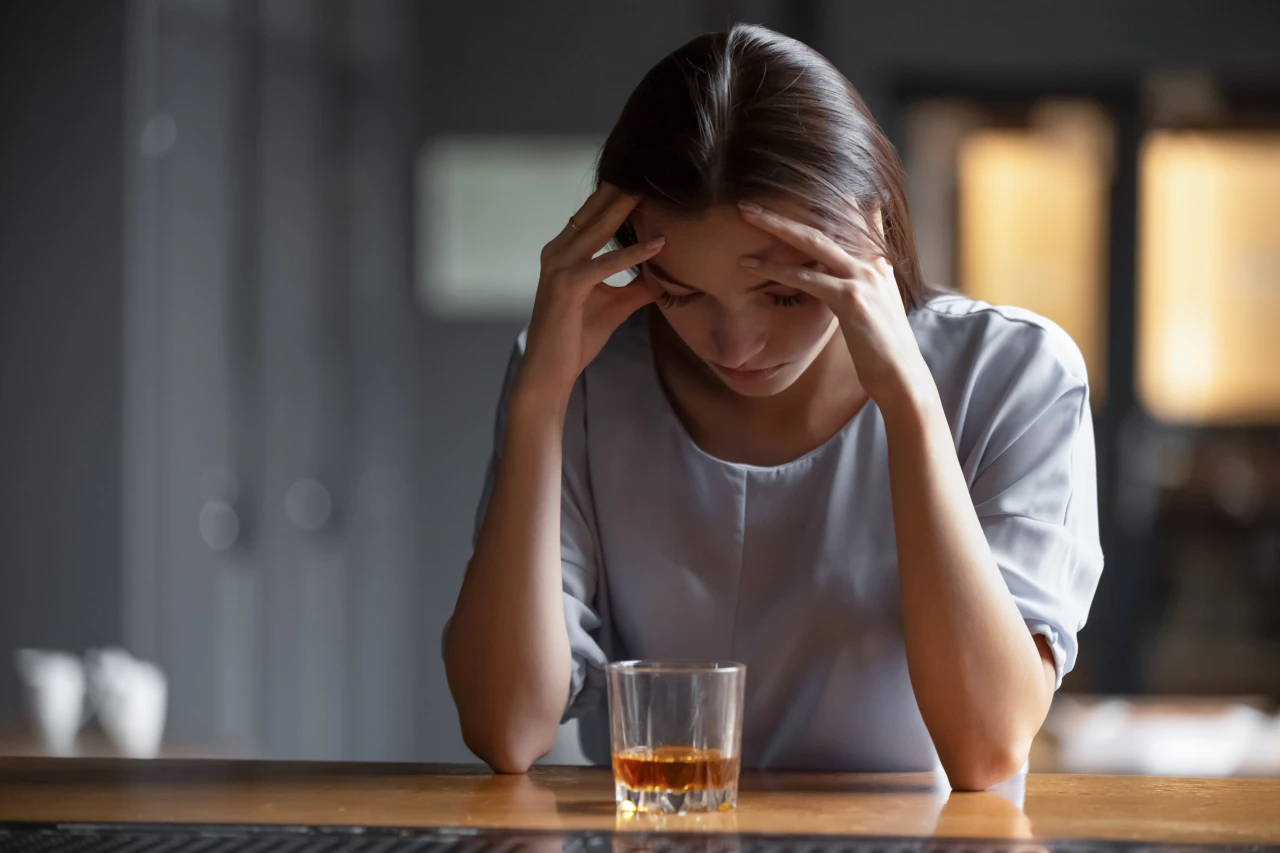 vrouw-grijze-blouse-en-glas-alcohol-houdt-hoofd-in-handen Vrouw met glas alcohol steunt hoofd in handen en is voorbeeld van hoe je verslaving herkent.