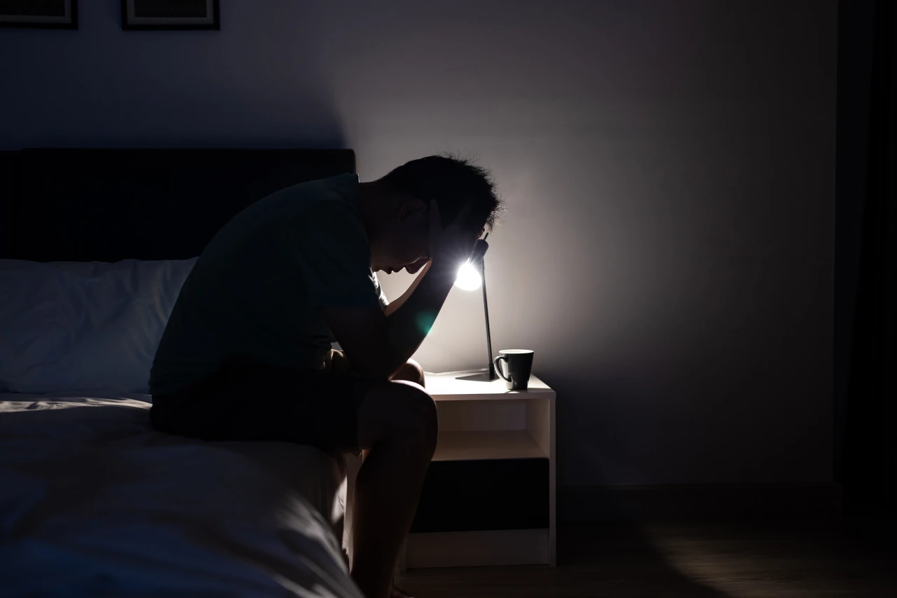 man-zit-op-rand-van-bed-met-hoofd-in-handen-en-alleen-nachtlampje-aan Man zit op rand van bed omdat hij niet kan slapen en houdt hoofd in handen van wanhoop.