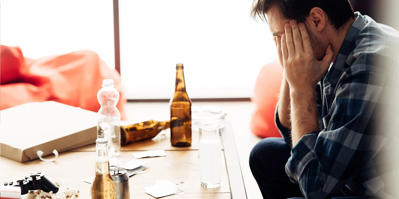 Een man zit met zijn handen voor zijn gezicht aan tafel met lege flessen alcohol en oogt uitgeput en gespannen.