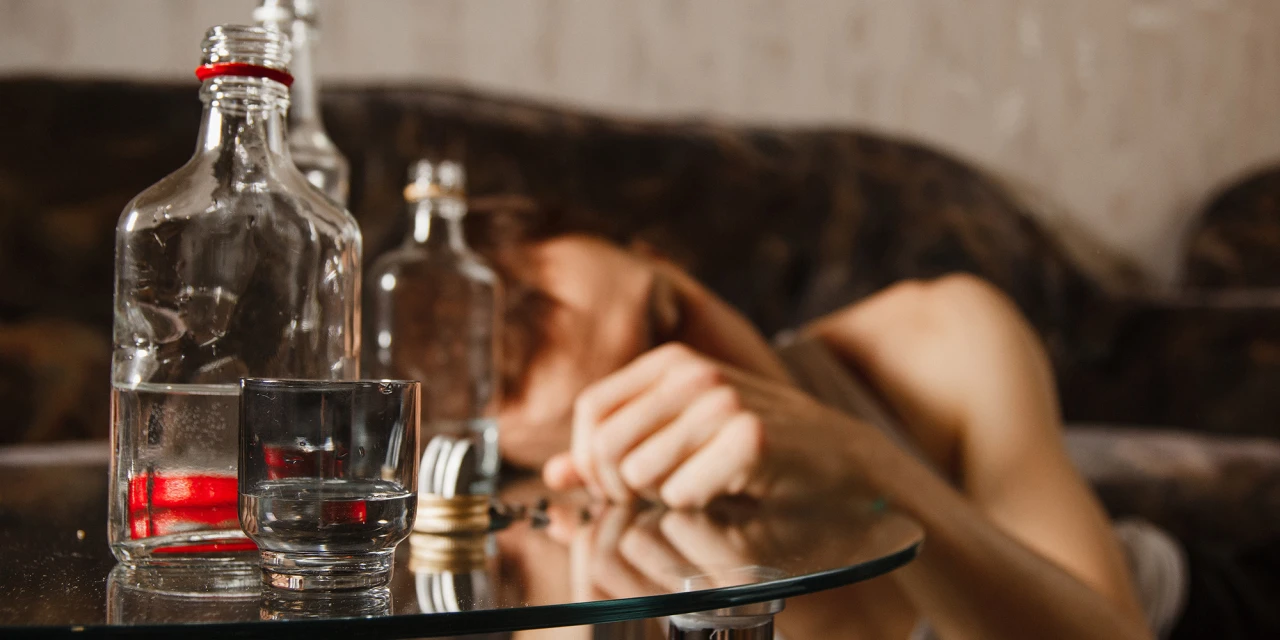 Een persoon ligt uitgeput op de bank terwijl lege alcoholflessen en een glas op tafel staan na overmatig alcoholgebruik.