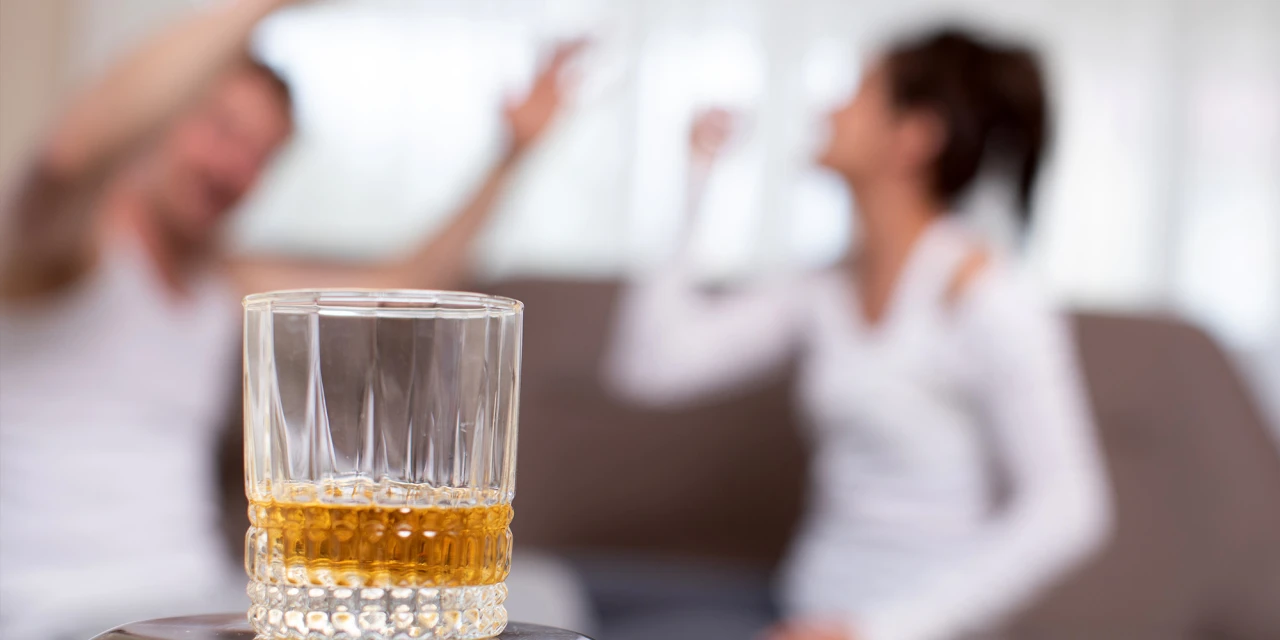 Een glas alcohol staat op de voorgrond terwijl een man en vrouw op de achtergrond ruzie maken.