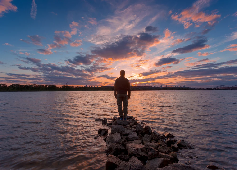 man-kijkt-naar-zonsopgang-aan-het-water Man staat op rotsen aan het water naar zonsopkomst of zonsondergang te kijken.